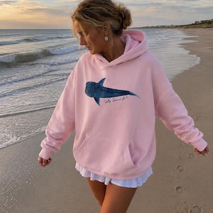 May include: A light pink hooded sweatshirt featuring a dark blue whale shark graphic on the chest with the text Salty Summer Art printed underneath. The garment includes a front kangaroo pocket, long sleeves with ribbed cuffs, and a hood.