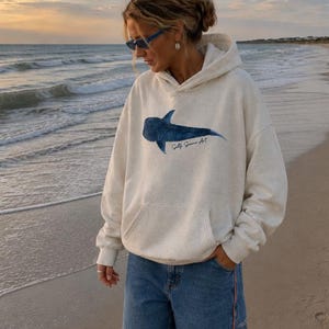 May include: An oatmeal-coloured hooded sweatshirt featuring a blue whale shark graphic on the chest with the text Salty Summer Art printed below it, paired with blue denim shorts that have a thin orange side stripe detail.
