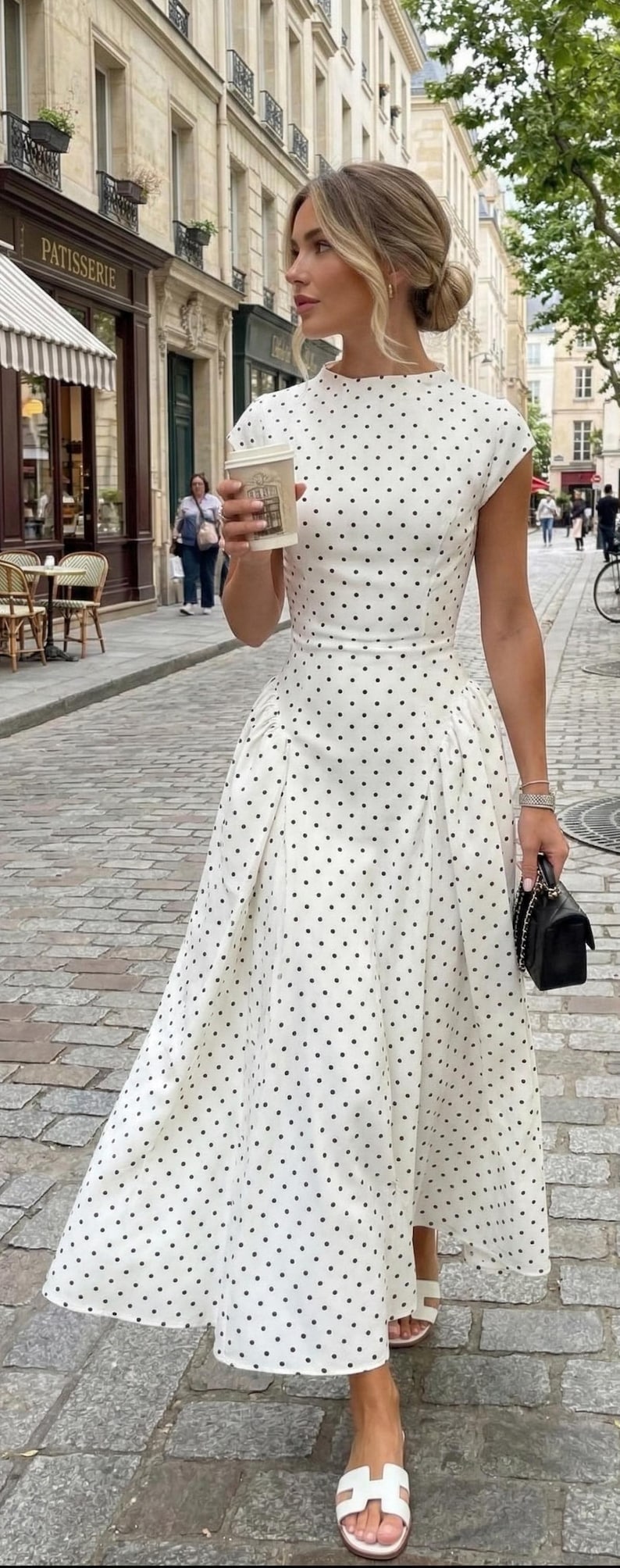 A woman stands on a cobblestone street wearing an elegant white and black polka dot maxi dress. The dress features a high neckline, cap sleeves, and a fitted waist with unique gathered detailing at the hips that flows into a voluminous ankle-length skirt. She holds a coffee cup and a small black handbag, pairing the look with white slide sandals. In the background, a charming patisserie and traditional urban architecture frame the scene.