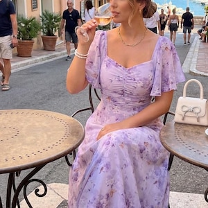 Peut inclure: Une femme porte une robe lavande à imprimé floral avec des manches courtes et un col carré. Elle est assise à une table de bistrot, tenant un verre de vin. Un petit sac à main blanc avec un nœud est posé sur la table. En arrière-plan, une rue européenne.