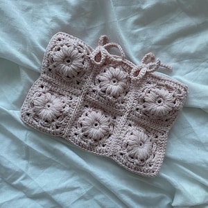 以下が含まれることがあります： 淡いピンク色の、花の模様の入ったかぎ針編みのバッグ。バッグは6つの正方形のパネルで構成されており、それぞれの中央に隆起した花があります。上端には2組の紐が付いています。バッグは柔らかい糸で作られています。