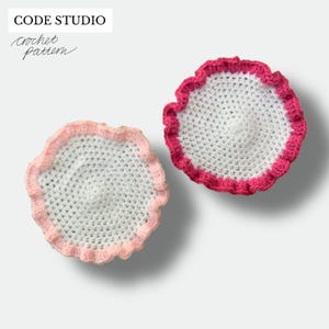 Op de afbeelding: Twee handgemaakte gehaakte onderzetters. Eén onderzetter heeft een wit midden met een roze gerimpelde rand. De andere onderzetter heeft een wit midden met een felroze gerimpelde rand. De tekst "CODE STUDIO Crochet Pattern" staat linksboven.