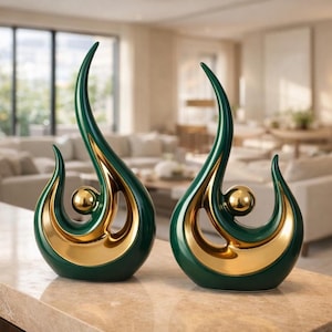 May include: Two decorative ceramic sculptures. Each sculpture features a teardrop shape with a gold interior and a dark green exterior. A gold sphere is centered within the design. The sculptures are displayed on a light-colored surface.
