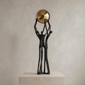 Op de afbeelding: Een bronzen sculptuur met drie figuren die een grote, reflecterende gouden bol ondersteunen. De figuren hebben langgerekte ledematen en staan op een vierkante, lichtgekleurde sokkel. De sculptuur heeft een getextureerde afwerking tegen een neutrale achtergrond.