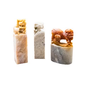 May include: Three carved stone seals in shades of beige, gray, and orange. Each seal features a detailed animal or mythical creature at the top. The seals are rectangular and oval-shaped, showcasing intricate craftsmanship.