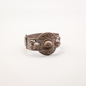 Puede incluir: Una pulsera de puño plateada con un diseño ornamentado. La pulsera presenta una pieza central circular grande con un patrón texturizado en relieve y detalles florales. Una delicada cadena conecta los dos lados.