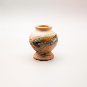 Puede incluir: Un pequeño jarrón decorativo de cerámica con base de pedestal. El jarrón presenta un diseño veteado en tonos marrón, naranja, turquesa y gris. La parte superior e inferior son de color beige liso. El jarrón mide unos 10 cm de alto.