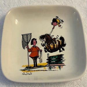 May include: A square white ceramic trinket dish featuring a colorful cartoon illustration by Thelwell. The graphic depicts a rider in an orange jacket holding a net, while a pony jumps over a green hedge, causing the rider to fall off.