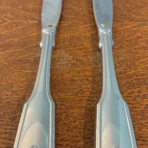 May include: Two silver-toned metal dinner knives featuring a smooth, tapered handle design. Each handle terminates in a decorative, raised shell motif at the base. The blades are straight with a slightly curved, pointed tip and a polished finish.