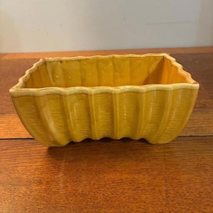 May include: A rectangular mustard yellow ceramic planter with a scalloped, wavy rim and vertical fluted detailing along the exterior walls. The vessel features a glossy finish and a hollow interior, with a slightly raised base design.