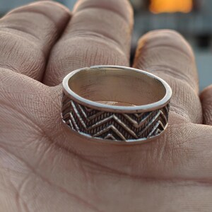 May include: A silver ring featuring a geometric chevron pattern. The wide band ring has a textured design. The ring is displayed on a hand.