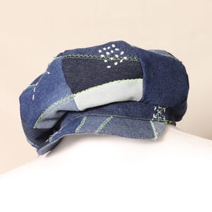 May include: A patchwork denim beret in various shades of blue, with accents of light green stitching. The beret features a unique design with visible seams and a textured appearance, showcasing a handcrafted aesthetic.