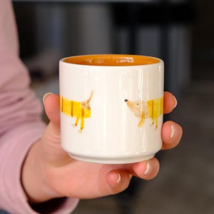 Könnte beinhalten: Eine weiße Keramiktasse mit orangefarbenem Innenraum, verziert mit Illustrationen von zwei Dackeln in gelben Pullovern. Die Tasse wird in der Hand einer Person gehalten.