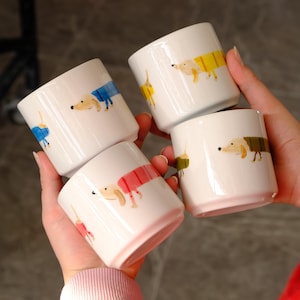Könnte beinhalten: Vier weiße Keramikbecher mit einem Dackel-Design. Jeder Becher zeigt einen unterschiedlich farbigen Mantel auf dem Hund: blau, gelb, rot und grün. Die Becher werden in den Händen einer Person gehalten.
