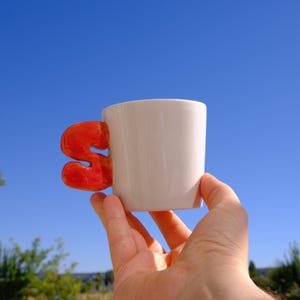 May include: A white ceramic mug featuring a glossy, bright orange handle shaped like the letter S. The mug has a smooth, cylindrical body and a vibrant, solid-colored handle that contrasts with the clean white finish of the cup's exterior surface.