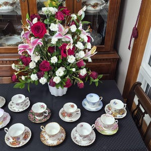May include: A display of vintage teacups and saucers with floral patterns. A large floral arrangement with red roses, white flowers, and pink lilies is in the center. The teacups are arranged on a black tablecloth, creating an elegant tea setting.