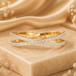 May include: A gold ring featuring a crossover design with two intersecting bands. Both bands are encrusted with small, clear, sparkling stones that cover the entire visible surface of the metal, creating a continuous shimmering effect across the ring.