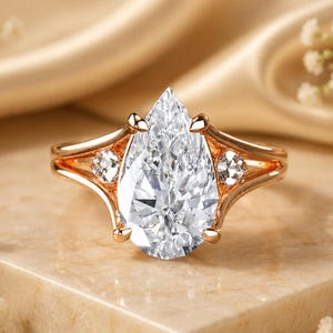 May include: A rose gold ring featuring a large pear-shaped clear center stone held by four prongs, flanked by two smaller round clear stones on each side of the split shank band, creating a symmetrical design with a polished metallic finish.