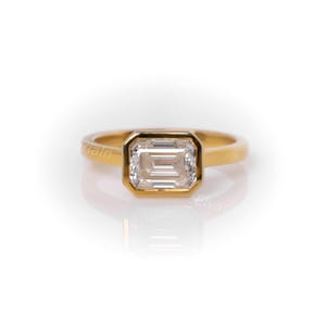 Könnte beinhalten: Ein goldener Ring mit einem rechteckigen, klaren Edelstein. Der Edelstein ist in einer Zargenfassung gefasst, und das Band ist ein schlichtes, poliertes Gold. Der Ring wird auf einer weißen Oberfläche mit einer leichten Reflexion gezeigt.