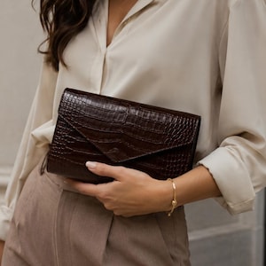 Puede incluir: Un bolso de mano tipo sobre en color marrón oscuro con un patrón texturizado de relieve de cocodrilo. El accesorio rectangular presenta un cierre de solapa en punta y un acabado brillante, combinado con una camisa beige y pantalones.