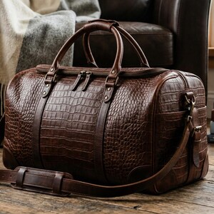 May include: A brown leather duffel bag with a crocodile texture, featuring a top handle and a shoulder strap. The bag is on a wooden floor, with books and a compass in the background. The bag measures approximately 50 cm long.