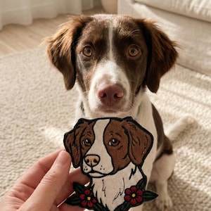Può includere: Una toppa per cani marrone e bianca con un cane abbinato sullo sfondo. La toppa presenta la faccia di un cane con pelo marrone e bianco, delineato in nero, e fiori rossi in basso. Il cane sullo sfondo ha una colorazione simile.