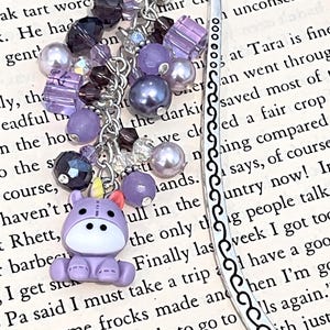 May include: A silver bookmark with a decorative charm. The charm includes a purple hippo, purple beads, and iridescent crystals. The bookmark is on a book with printed text.