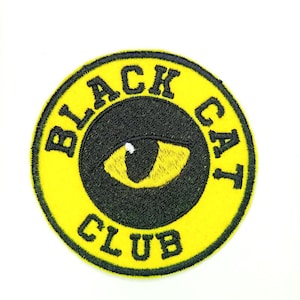 Puede incluir: Un parche redondo bordado, amarillo y negro, con el texto "BLACK CAT CLUB" que rodea un ojo de gato negro. El ojo es amarillo con un contorno y una pupila negros. El parche está sobre un fondo blanco.
