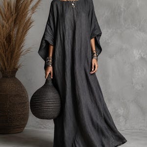 Könnte beinhalten: Ein bodenlanges Kaftan-Kleid in Anthrazit mit weiten, lockeren Ärmeln und einer strukturierten Stoffoberfläche. Das Kleidungsstück hat einen einfachen Rundhalsausschnitt und eine fließende, lockere Silhouette, kombiniert mit dunkelbraunen flachen Sandalen.