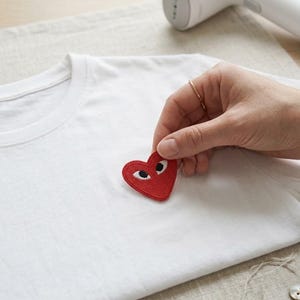 May include: A white t-shirt featuring a red heart-shaped patch with two white eyes and black pupils. The patch is being positioned on the chest area of the fabric, with a white handheld garment steamer visible in the upper portion of the frame.