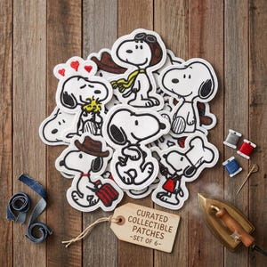 Könnte beinhalten: Eine Sammlung von gestickten Snoopy-Aufnähern in verschiedenen Outfits und Posen, darunter ein Pilot, ein Koch und ein Reisender. Ein Holzanhänger mit dem Text CURATED COLLECTIBLE PATCHES -SET OF 6- liegt neben Garnrollen und einem Bügeleisen.