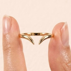 May include: A polished gold ring featuring an open design with two tapered, curved points that meet in the center. The band is smooth and rounded, showcasing a reflective metallic finish that catches the light across its surface.