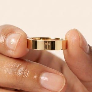 May include: A polished gold-tone band ring featuring a small, centred cross engraving. The ring has a flat, smooth surface with a reflective metallic finish and a simple, uniform width throughout the entire circumference of the circular band.