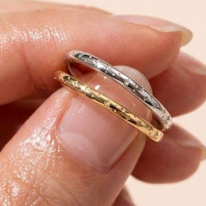 May include: Two thin, stackable rings, one in a polished silver tone and one in a polished gold tone. Each band features a delicate, repeating engraved star pattern encircling the entire circumference of the metal surface.