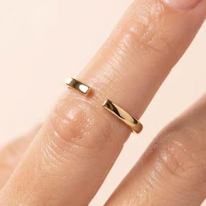 Peut inclure: Une bague ouverte en or avec une finition lisse et polie. L'anneau présente un espace entre les deux extrémités, créant un aspect propre et épuré. La bague est portée sur un doigt, mettant en valeur sa surface métallique simple et réfléchissante.