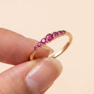 Pode incluir: Um anel de ouro com uma fileira de sete pedras preciosas rosa vibrantes. A banda do anel é fina, e as pedras preciosas são colocadas próximas umas das outras, criando uma linha contínua de cor. O anel é segurado contra um fundo macio e neutro.
