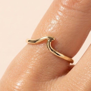 Puede incluir: Un anillo de oro pulido que presenta un elemento de diseño único en forma de onda suave en la banda. El anillo tiene un perfil continuo y redondeado con un acabado metálico reflectante, rodeando el dedo con una estética simple, fluida y minimalista.
