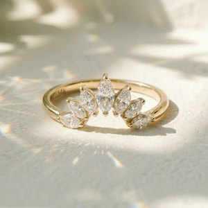 Puede incluir: Un anillo de oro con una fila curva de diamantes talla marquesa. La banda del anillo es delgada y pulida, y los diamantes están engastados en una delicada disposición en forma de corona. El anillo se muestra sobre una superficie blanca.