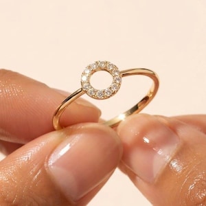 Pode incluir: Um anel de ouro com uma peça central circular adornada com uma série de pequenas pedras claras de corte redondo. A banda é fina e polida, apresentando um acabamento metálico que reflete a luz, criando uma aparência brilhante e uniforme.