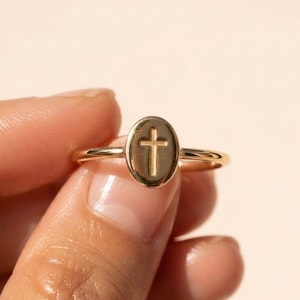 Könnte beinhalten: Ein goldener Ring mit einer polierten ovalen Oberfläche, in deren Mitte ein vertieftes Kreuzsymbol eingearbeitet ist. Der Ring ist dünn und kreisförmig gestaltet und weist durchgehend eine einheitliche metallische Oberfläche auf.
