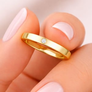 May include: A gold band ring with a single, small, round, clear gemstone. The ring is held up against a soft pink background. The ring is a simple, elegant design.