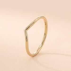 May include: A gold-coloured ring with a V-shaped design. The ring is made of a shiny, smooth metal and has a minimalist style. The ring is set against a soft, pink background.