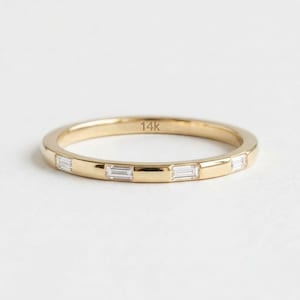 May include: A gold band ring featuring four evenly spaced baguette-cut clear stones set into the metal. The interior of the band is clearly stamped with the text 14k, indicating the gold purity of this polished yellow gold jewellery piece.