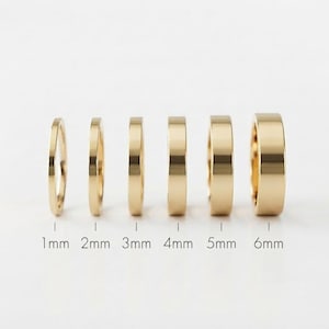 Può includere: Sei anelli color oro disposti in fila, che aumentano di larghezza da sinistra a destra. Gli anelli sono etichettati con le rispettive larghezze di 1mm, 2mm, 3mm, 4mm, 5mm e 6mm, indicando chiaramente la progressione delle dimensioni delle fasce lisce.