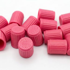 Puede incluir: Una colección de pequeños dedales cilíndricos de color rosa con un exterior texturizado y acanalado, y una superficie superior plana con relieve. Estos accesorios de costura están dispuestos en un montón, mostrando su color rosa uniforme.