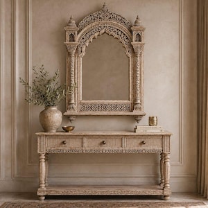 May include: A light-toned wooden console table with three drawers and a lower shelf, paired with an ornate, arched wall mirror featuring intricate carved detailing. A textured ceramic vase with greenery sits on the table surface.