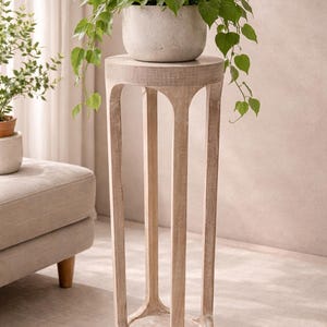 Puede incluir: Un soporte de planta alto de madera clara con una parte superior redonda y cuatro patas arqueadas. Una maceta redonda gris moteada llena de plantas verdes y frondosas se encuentra encima. Las enredaderas de la planta caen por los lados del soporte.