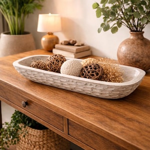 May include: A wooden, rectangular decorative bowl filled with decorative spheres and dried grasses. The bowl is light gray and sits on a dark brown wooden table. A lamp and potted plants are in the background.