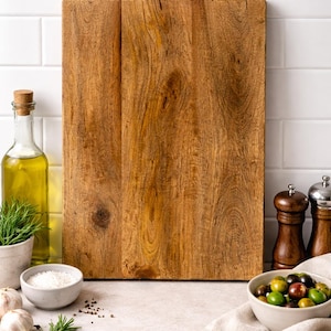 Könnte beinhalten: Ein rechteckiges Holzschneidebrett mit sichtbarer Holzmaserung. Eine Flasche Olivenöl, eine kleine Schüssel Salz, Knoblauch, Rosmarin, Oliven und Salz- und Pfeffermühlen sind um das Brett angeordnet, was auf ein kulinarisches Thema hindeutet.