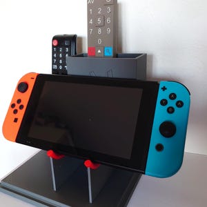 Peut inclure: Un support gris imprimé en 3D tenant une console Nintendo Switch noire avec des manettes orange et bleues. Le support contient également deux télécommandes et des supports rouges. Une télécommande grise est à l'arrière-plan.